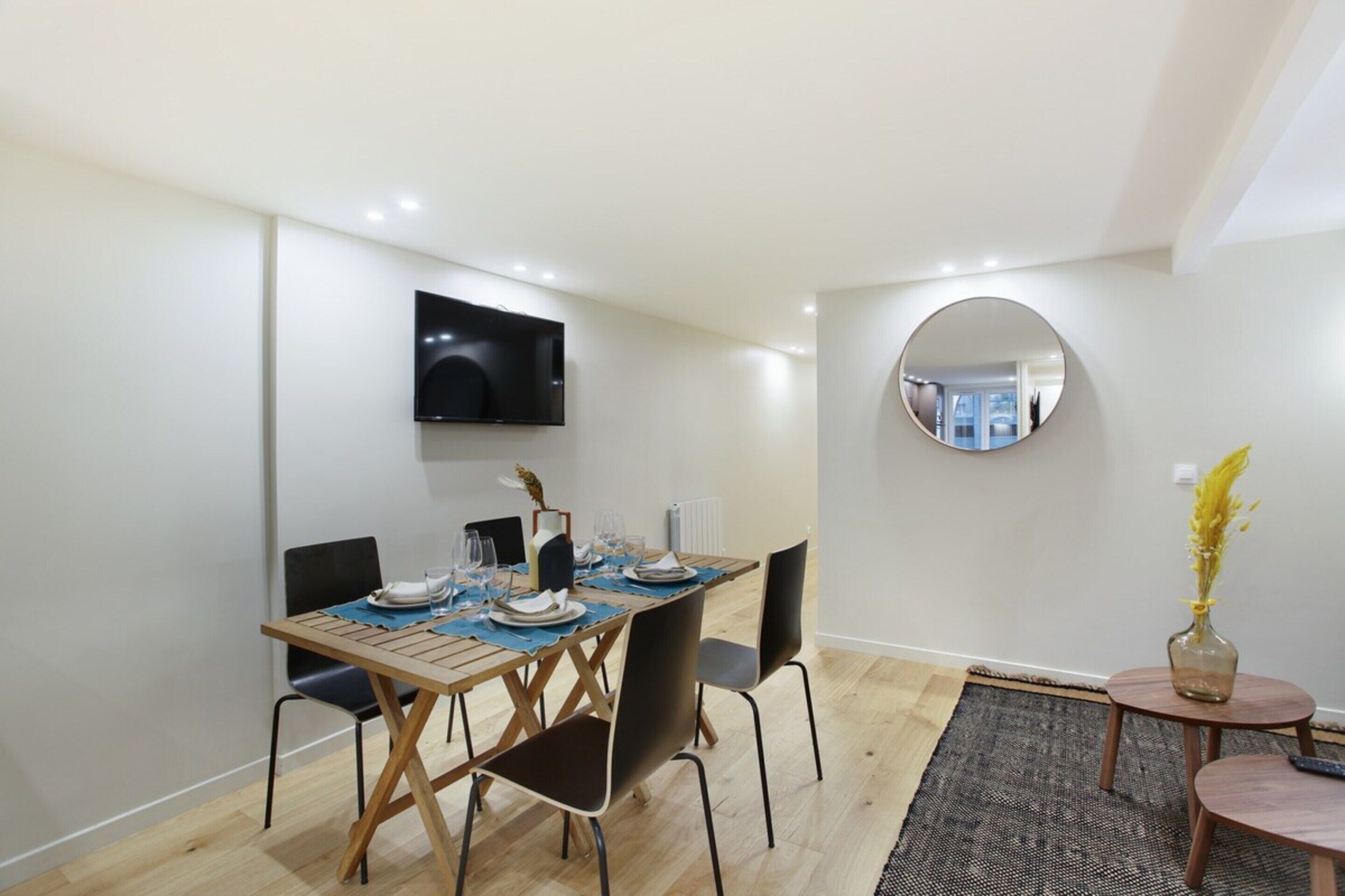 A dining area with a wooden table set for four, a flat-screen TV mounted on the wall, and a round mirror on the wall.