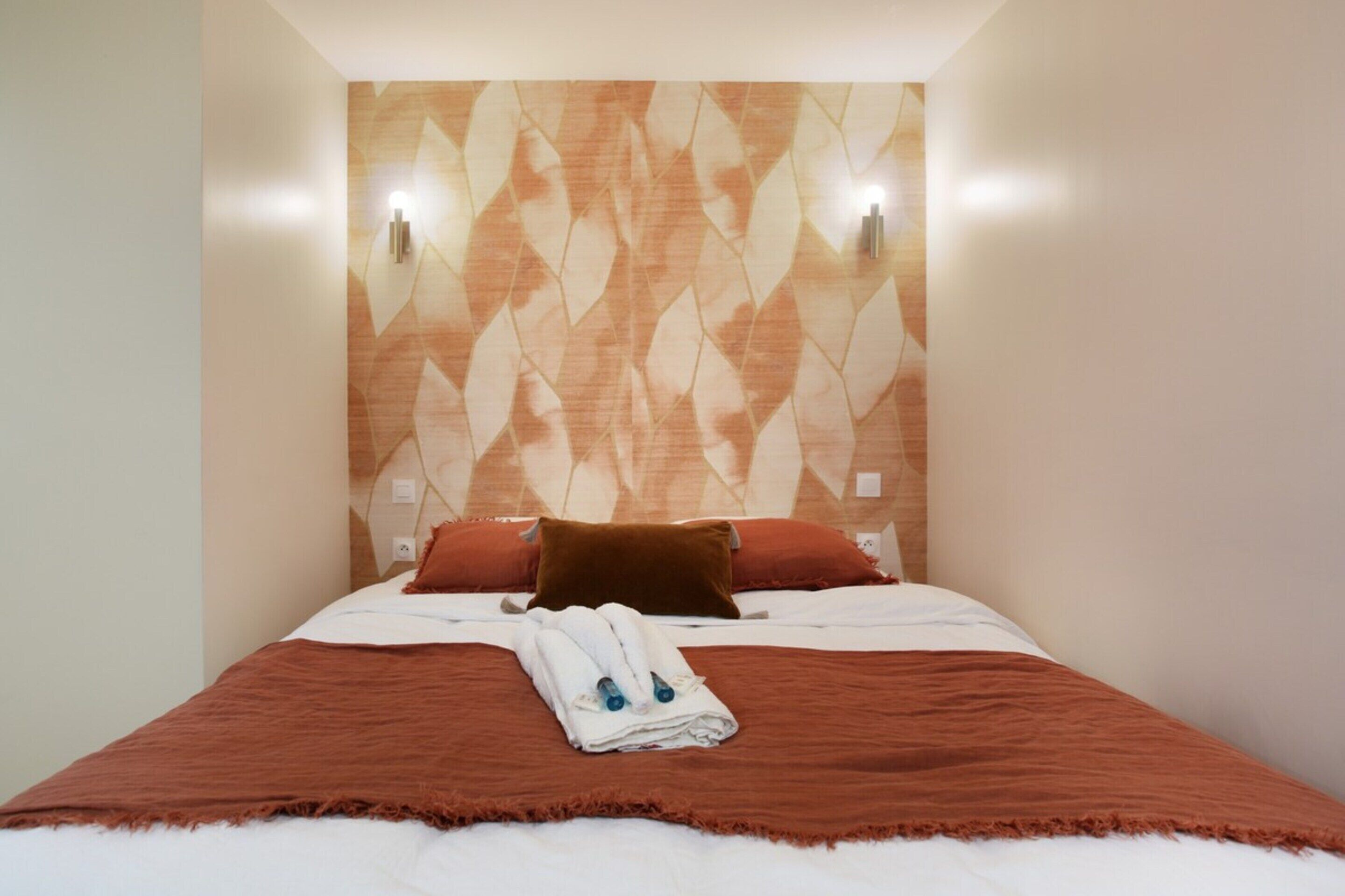 A neatly made bed with a brown blanket and pillows, a folded towel, and two wall-mounted lights.
