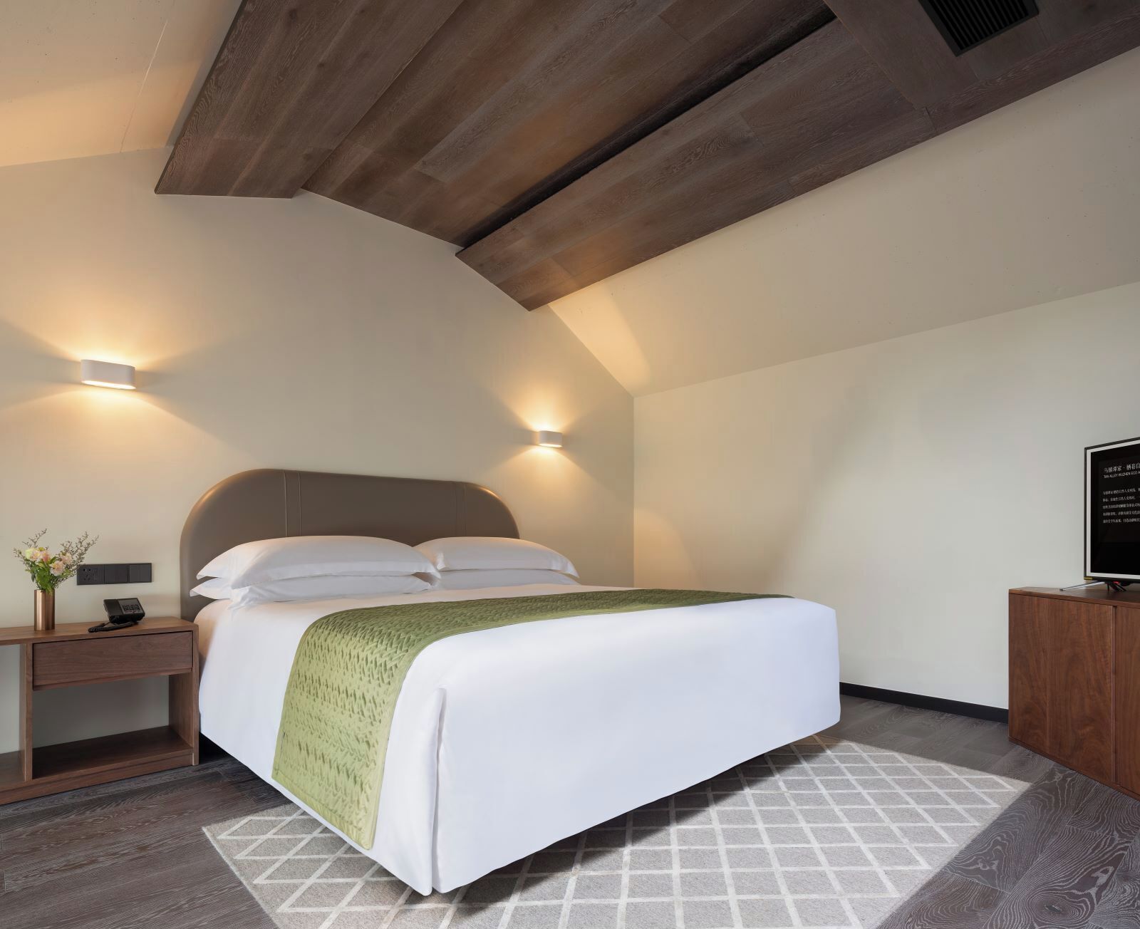 Ein modernes Hotelzimmer mit einem großen Bett, Nachttischlampen, einem Nachttisch, einem Holzkasten und einem an der Wand befestigten Fernseher.