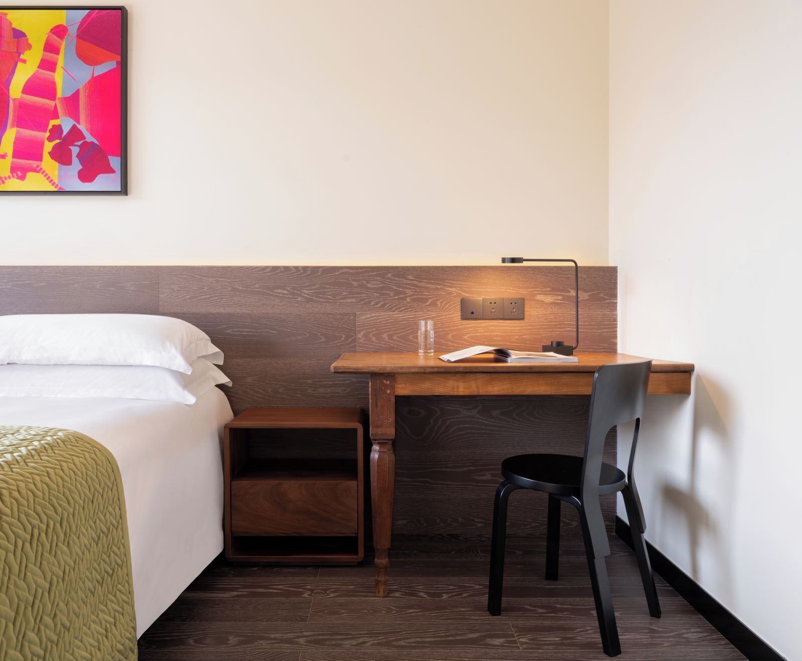 Habitació d'hotel amb un llit, escriptori amb cadira, una petita prestatgeria i una làmpada penjada a la paret.