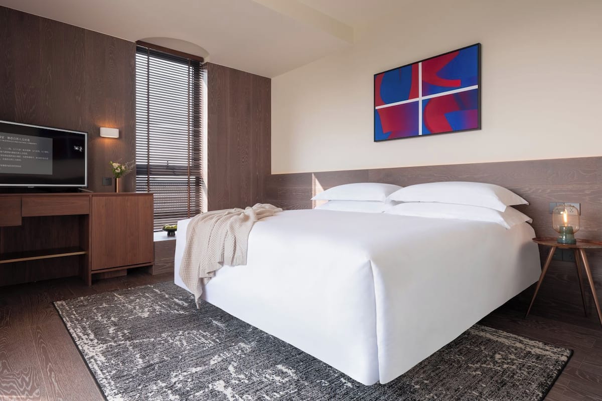 A modern hotel room with a large bed, a flat-screen TV, wooden paneling, and a colorful abstract painting.