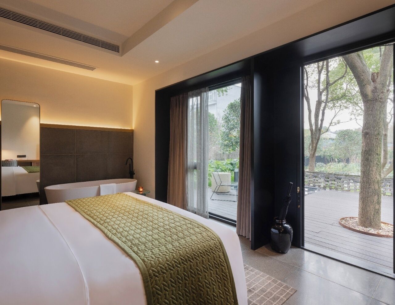 Une chambre d’hôtel moderne dotée d’un grand lit, d’un coin salon avec un fauteuil et une petite table, et offrant une vue sur l’extérieur grâce à une porte coulissante.