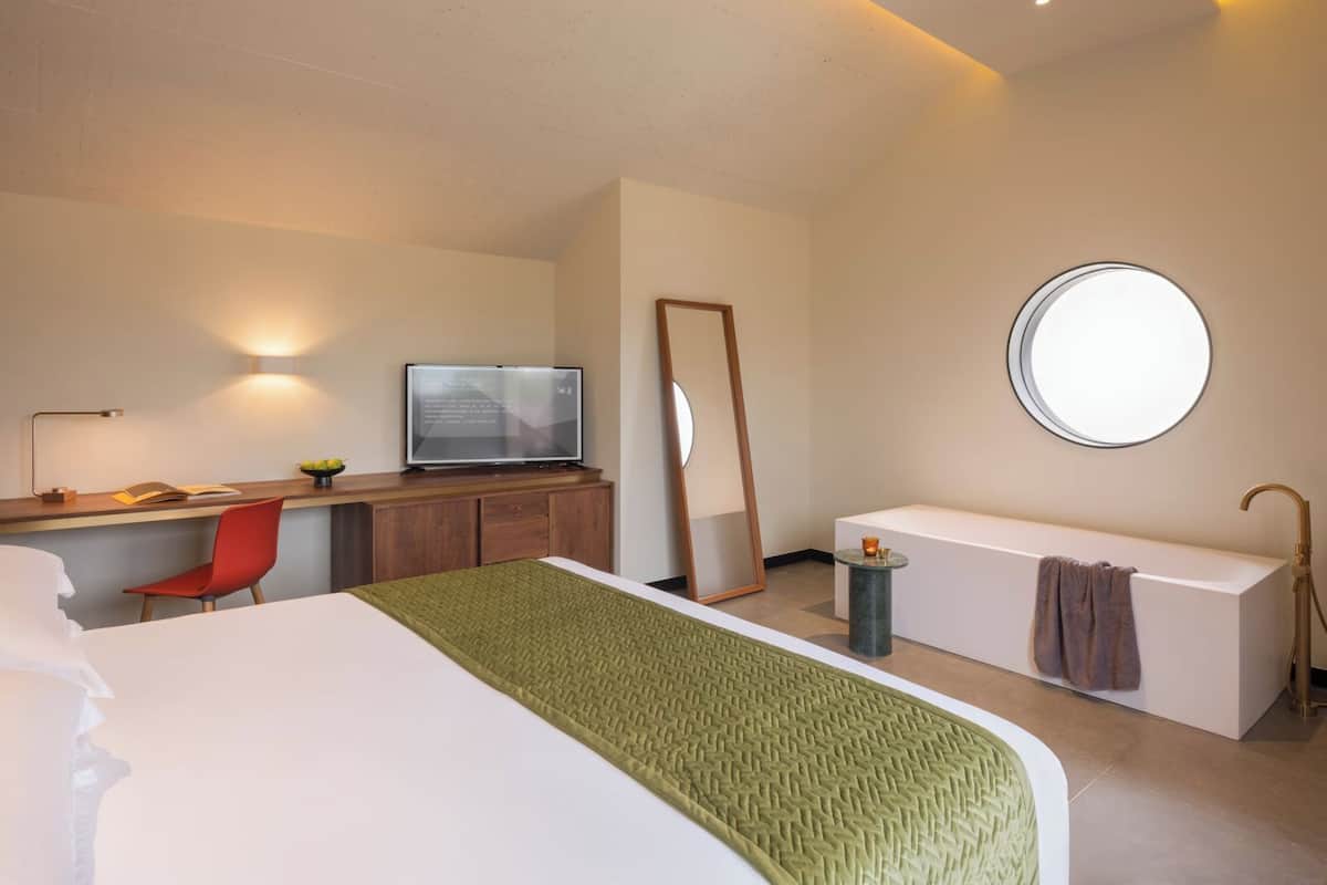 A modern hotel room with a bed, a desk with a laptop, a red chair, a round mirror, and a round window.