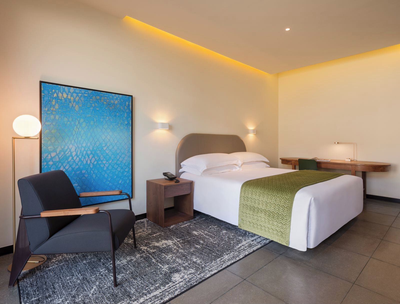 A hotel room with a bed, a desk, a chair, a nightstand, a lamp, and a large abstract painting on the wall.