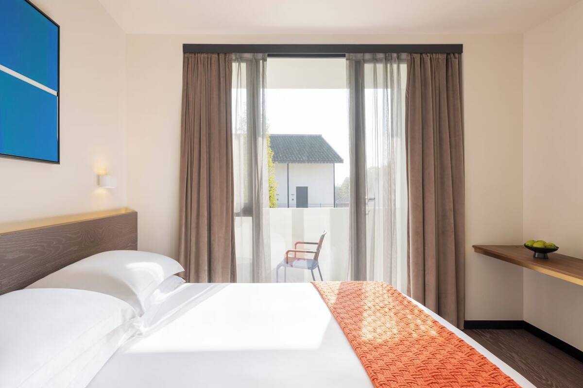A hotel room with a bed, a chair, a small table with a bowl of fruit, and a view of a building outside.