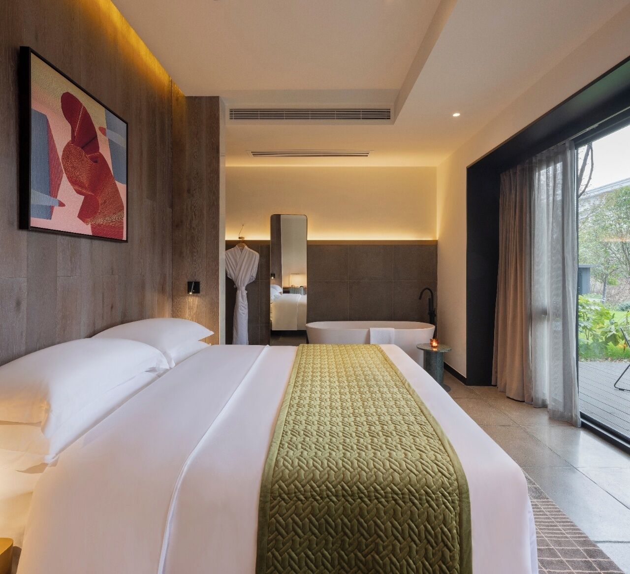 A hotel room with a large bed, a bathroom with a bathtub, and a view of greenery outside.