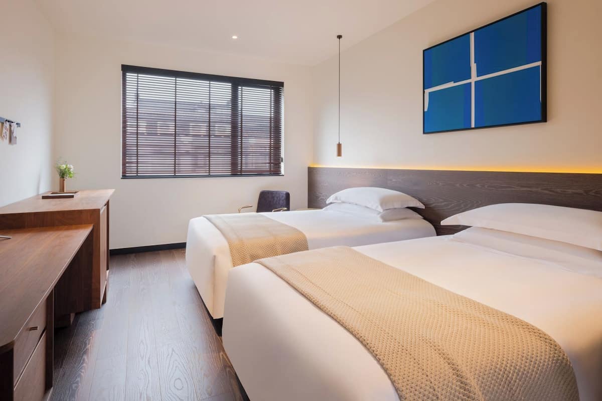 A hotel room with two beds, a desk, a window with blinds, and a blue wall art piece.