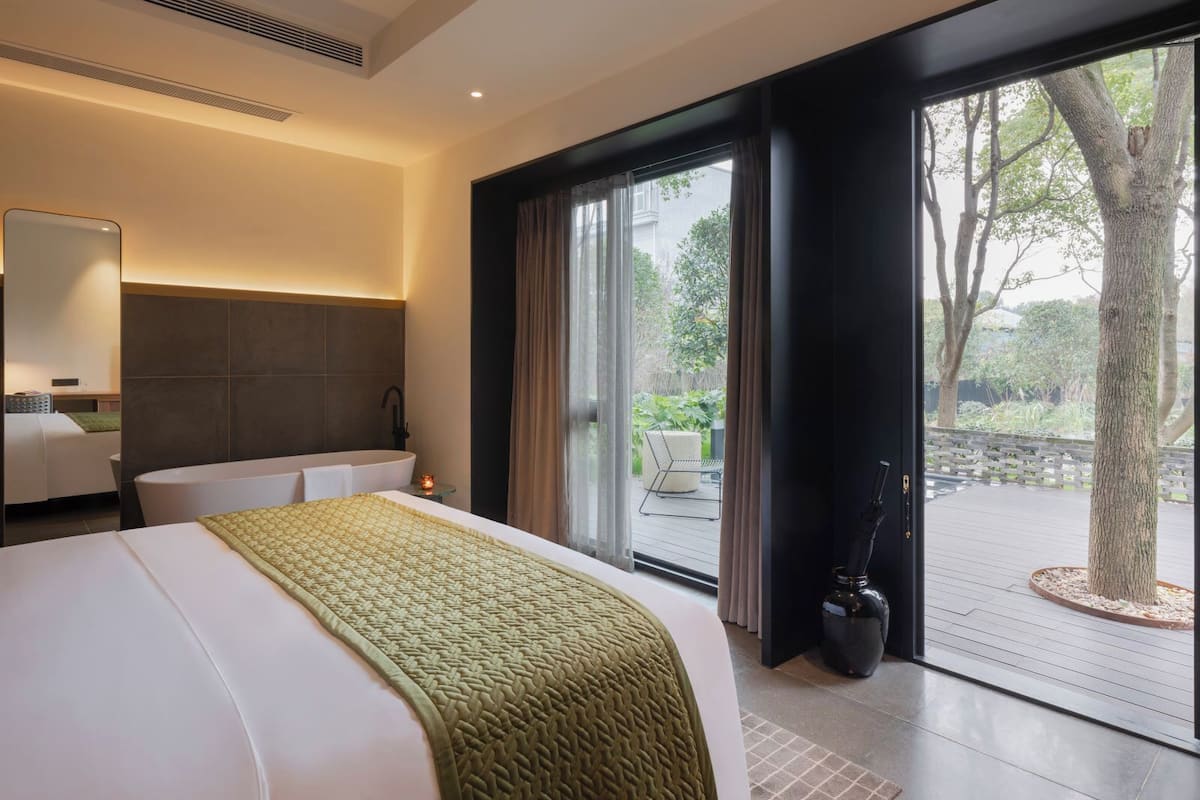 A modern hotel room with a large bed, a bathroom with a bathtub, and a view of a garden through a sliding door.