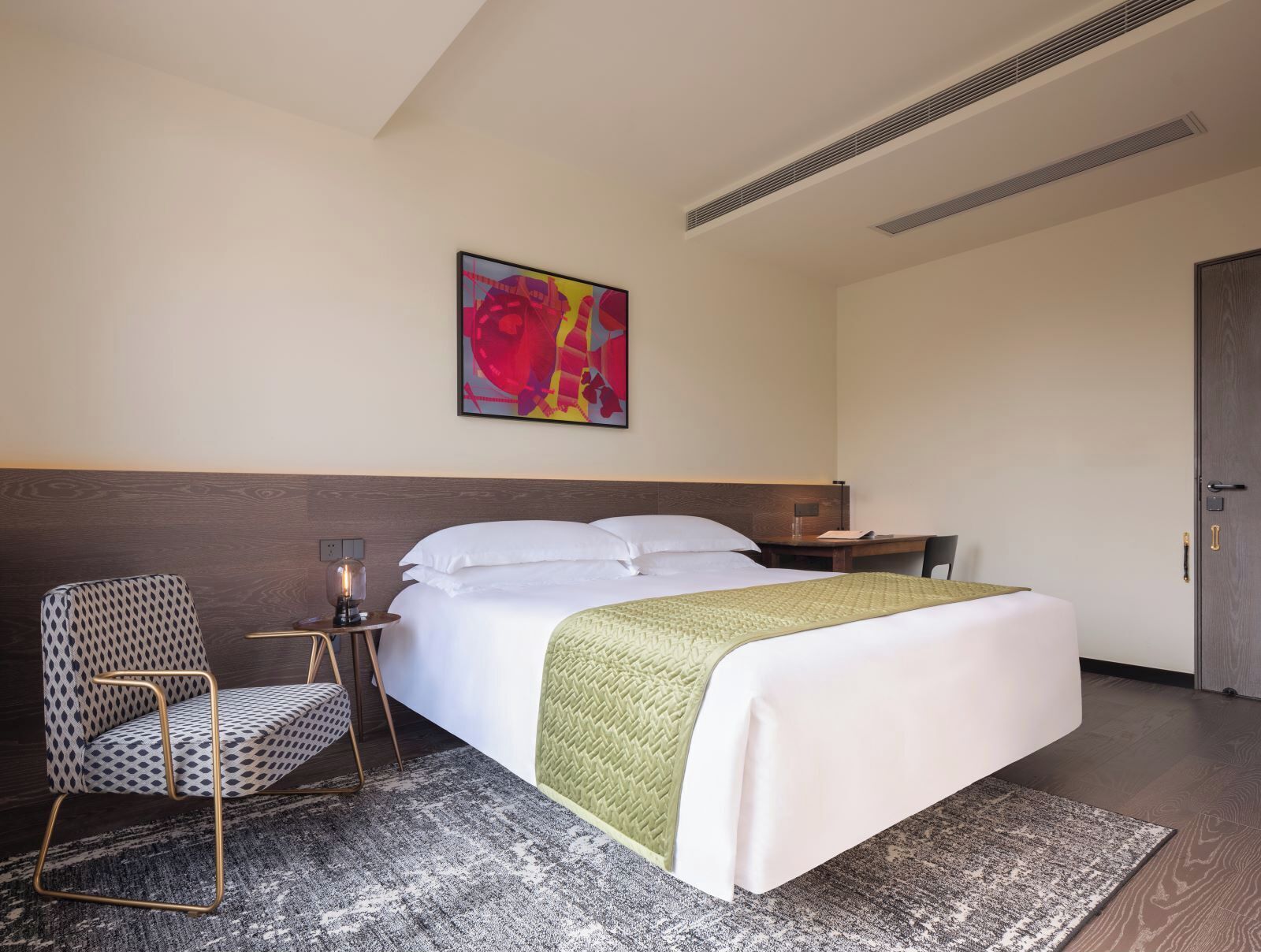 A hotel room with a bed, a chair, a desk, and a wall-mounted artwork.