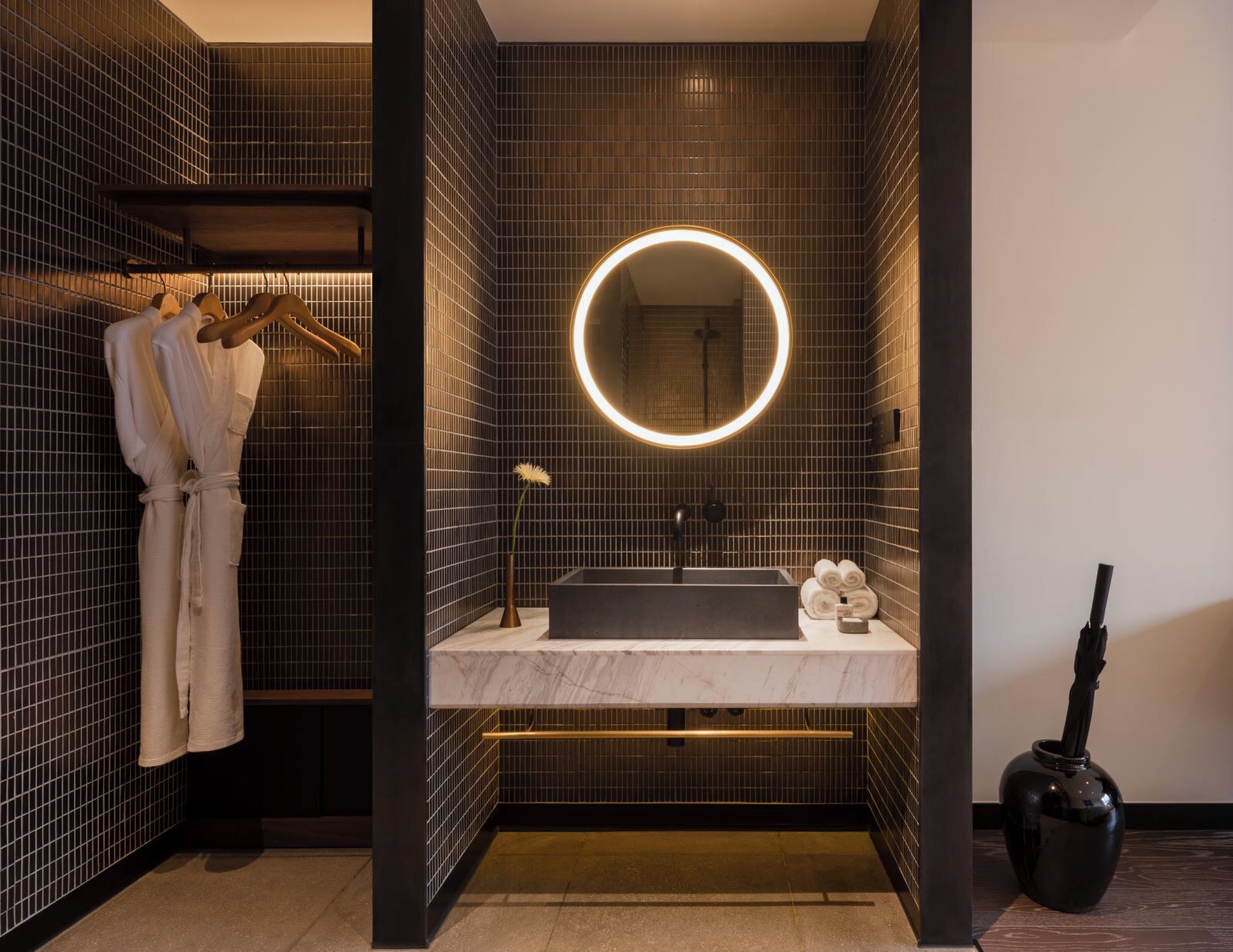 Un baño moderno con pared de azulejos oscuros, un espejo redondo con iluminación perimetral, un lavabo de mármol y un perchero con batas.