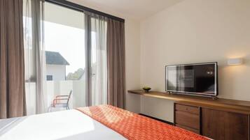 Hotellihuone, jossa on sänky, puinen hylly, jonka päällä on televisio, ikkuna, jossa on verhot, ja näkymä ulkona olevaan rakennukseen.