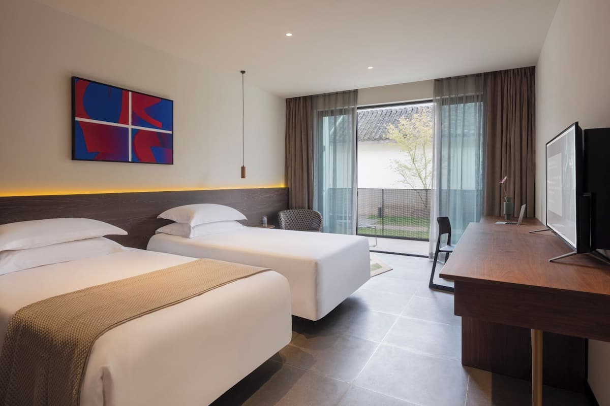 A hotel room with two beds, a desk with a computer, a TV, and a large window with curtains.