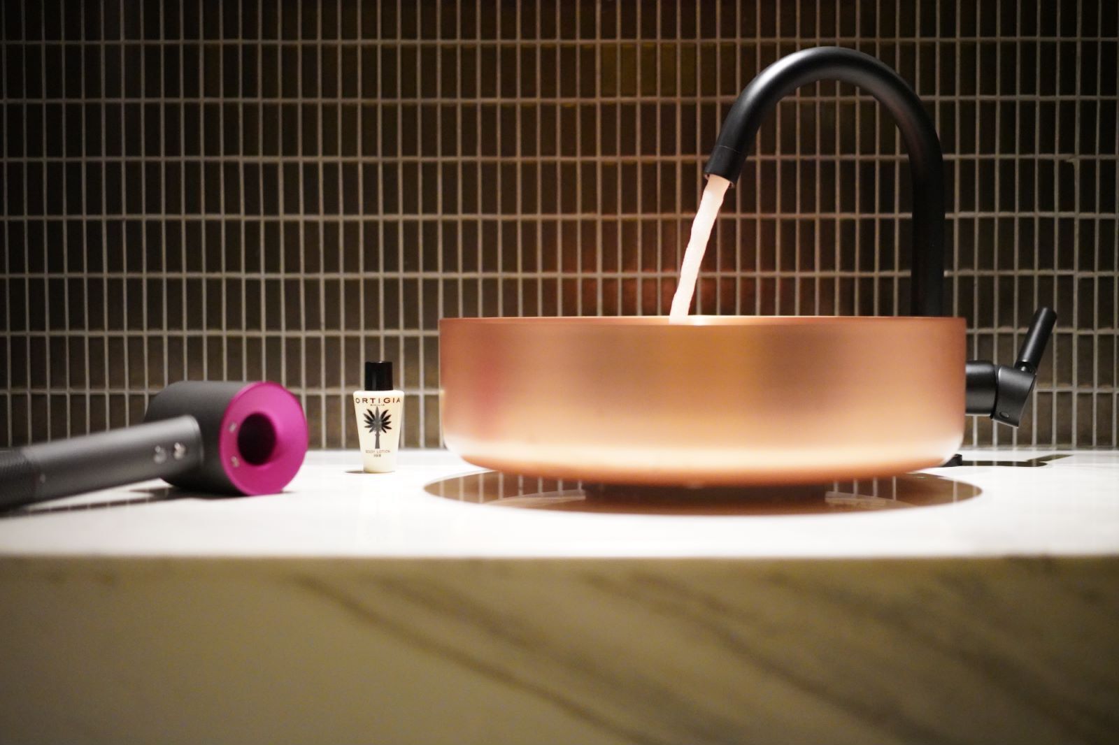 Una tetera color cobre sobre una encimera blanca, con un asa negra, un objeto rosa y una botella pequeña con la etiqueta "ORTIGIA" al fondo.