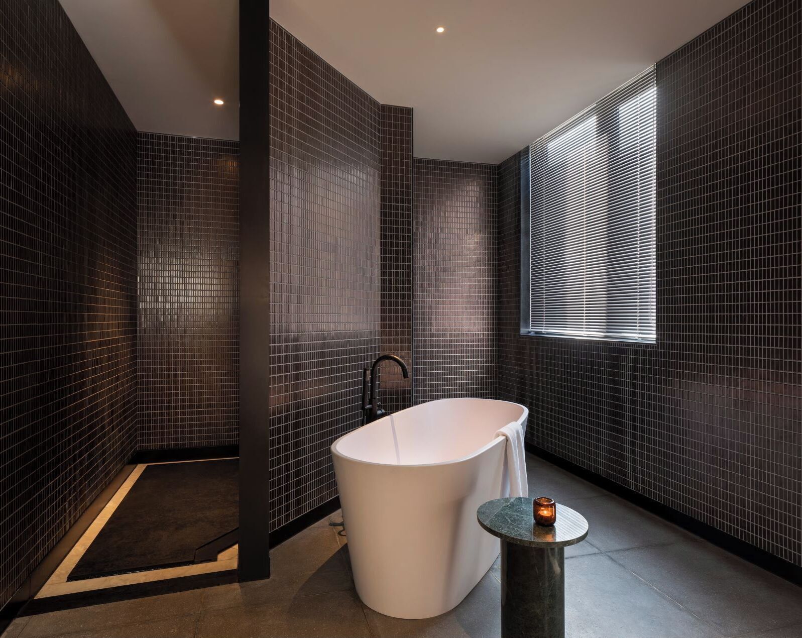 A modern bathroom with a freestanding bathtub, a small round table with a candle, and a window with blinds.