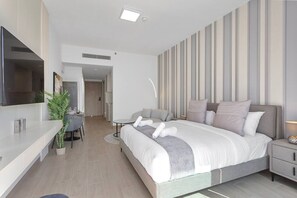 A modern hotel room with a large bed, bedside tables, a TV, and a dining area with a plant. - Perfect Getaway modern Studio in Luma 22 - JVC (Dubai)