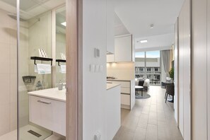 A modern hotel room with a glass shower, a white bathroom, a kitchenette, and a dining area with a view.