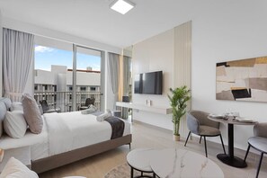 A modern hotel room with a large bed, a flat-screen TV, a dining area with a table and chairs, and a balcony with a view of the city.