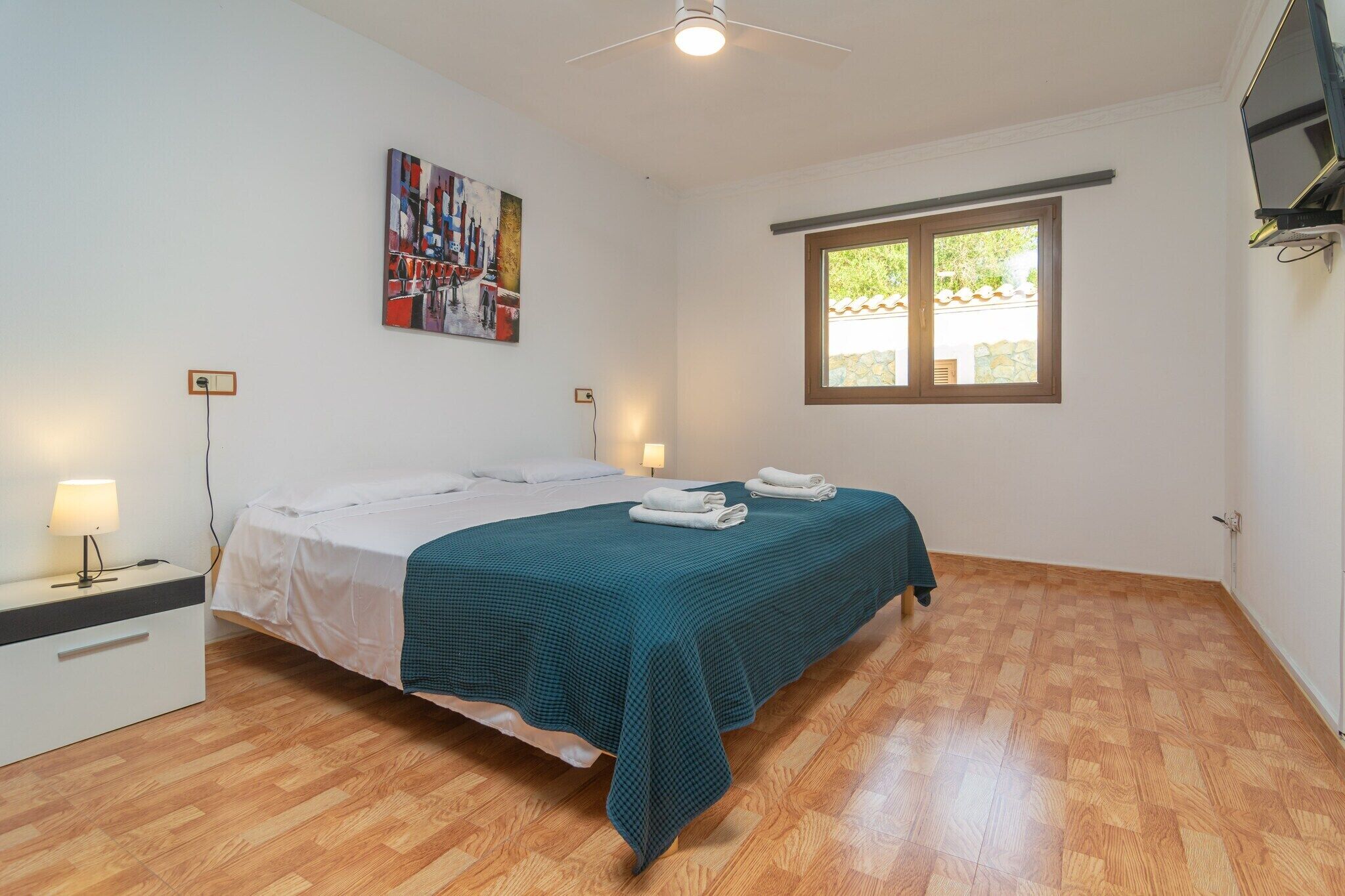 A bedroom with a bed, a nightstand, a lamp, a painting, a window, and a ceiling fan.