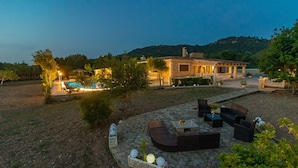 A stone patio with outdoor furniture, a swimming pool, and a building with a lighted entrance.