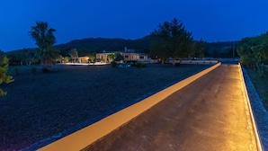 A paved pathway leading to a building with a lighted entrance, surrounded by trees and a hilly landscape.