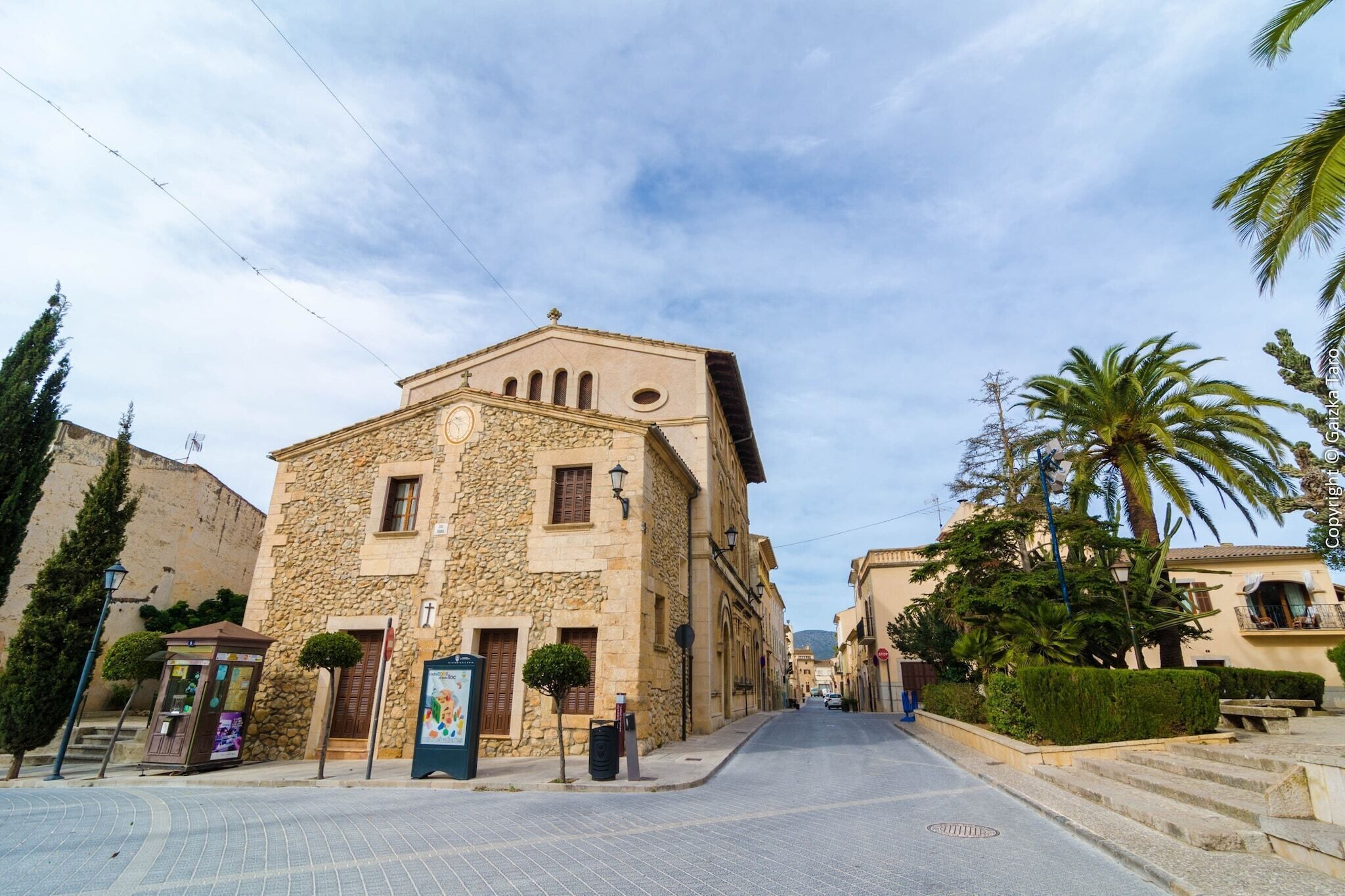 A stone building with a clock tower, a kiosk, and palm trees.
