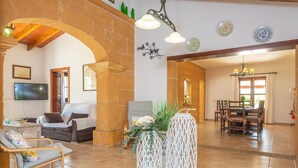 A spacious living room with a stone archway, a dining area with a wooden table and chairs, and a TV mounted on the wall. - 3 C'an Boto - Villa With Private Pool In Manacor (Manacor, Illes Balears)