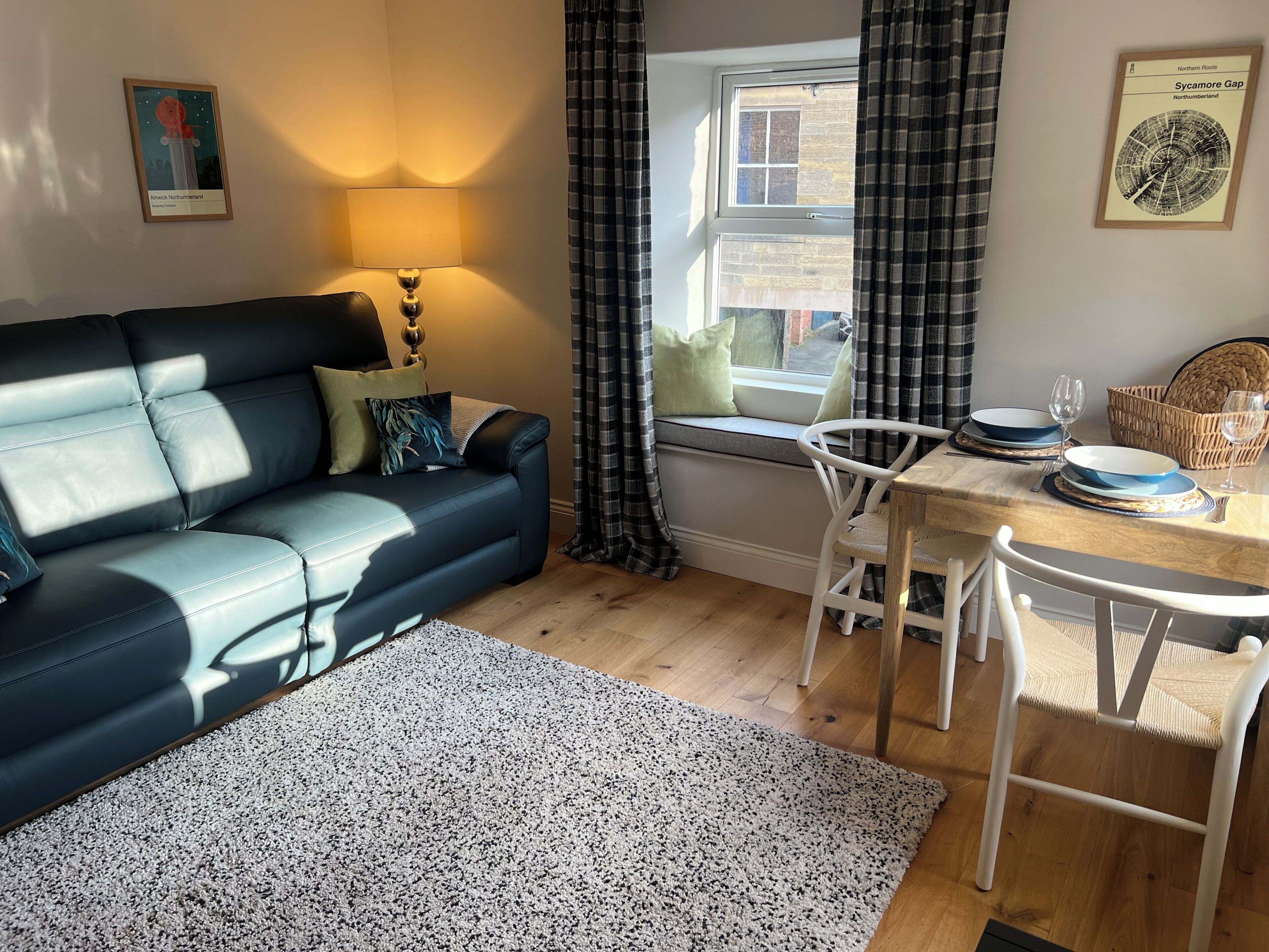 Un salon chaleureux avec un canapé noir, une petite table à manger entourée de chaises blanches, une fenêtre avec des rideaux et une lampe.