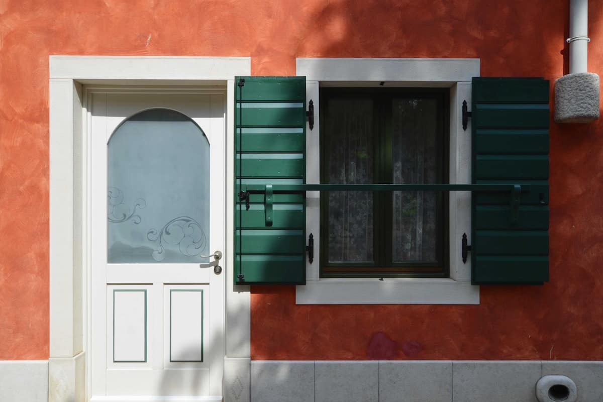 Facciata di un edificio con una porta bianca, persiane verdi, una finestra che si affaccia su alberi e un muro arancione con una superficie strutturata.