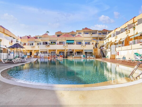 A pool area with lounge chairs and umbrellas, surrounded by multi-story buildings with balconies.