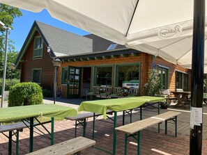 Een houten gebouw met een groene deur en ramen, buitentafels met groene tafelkleden, en een overdekt terras met een witte luifel.
