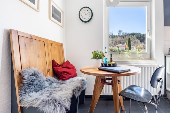 A room with a wooden headboard bed, a fluffy grey throw, a round wooden table with a plant, a black chair, and a view of a landscape through a window.