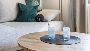 Pyöreä puinen pöytä, jonka päällä on kaksi lasillista vettä kudotun alustan päällä, sohva koristetyynyineen ja taustalla oleva lamppu.