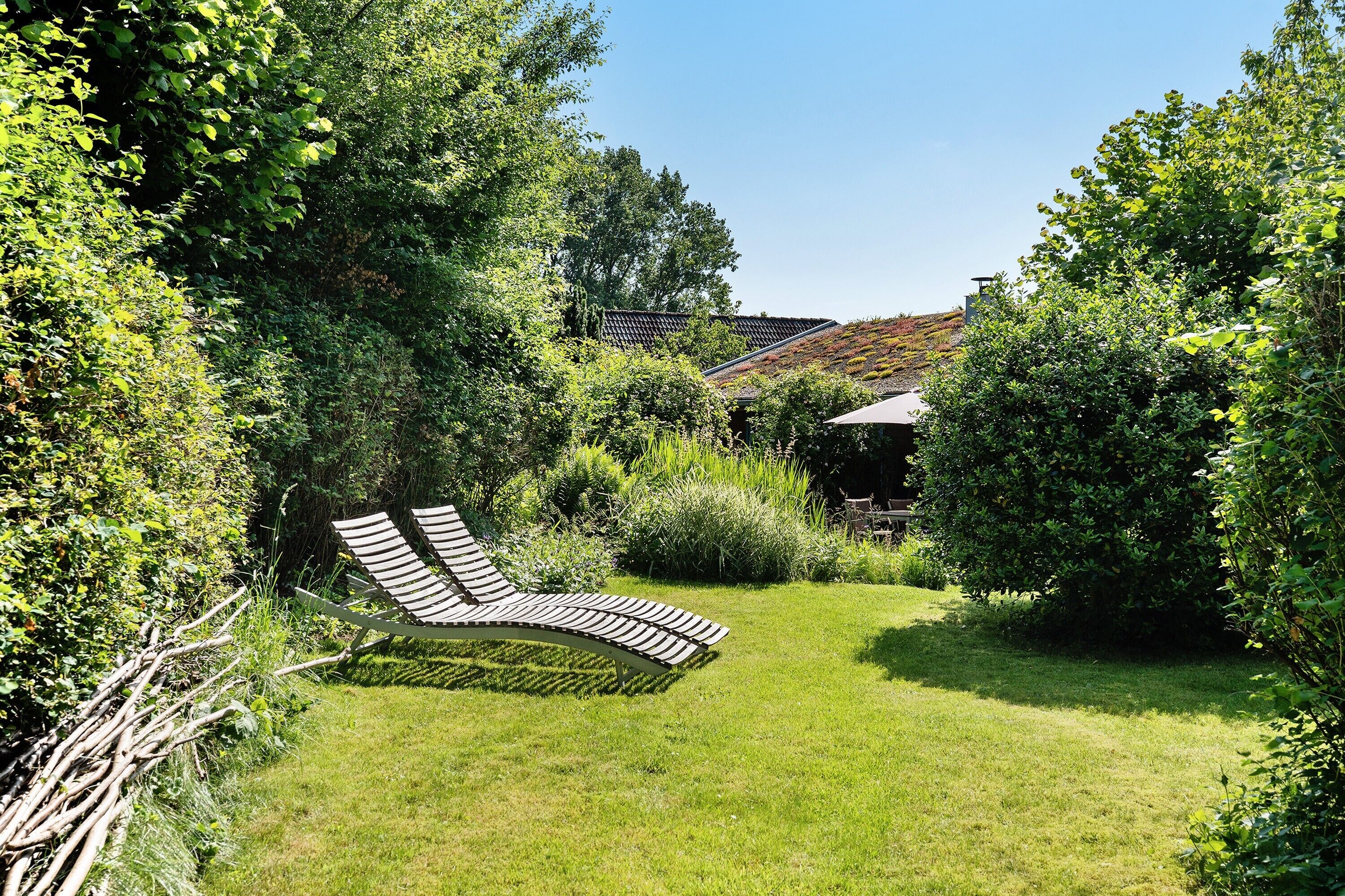 Ein Garten mit einer gebogenen Liege, üppiger Bepflanzung und einem Gebäude mit geneigtem Dach im Hintergrund.