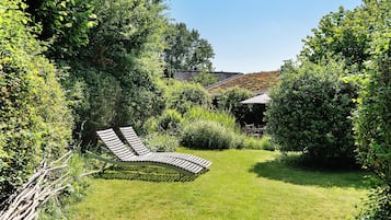Un jardín con una silla lounge curvada, follaje denso y un edificio con techo inclinado al fondo.