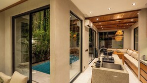 A modern interior with a glass door leading to a pool, a kitchen area with a sink, and a seating area with a sofa and table. - Nanna Villa Unit 1 by Betterplace (Canggu)
