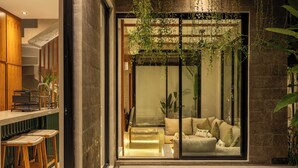 A modern kitchen with wooden stools, a built-in bar, and a glass-enclosed living area with a sofa and cushions. - Nanna Villa Unit 1 by Betterplace (Canggu)