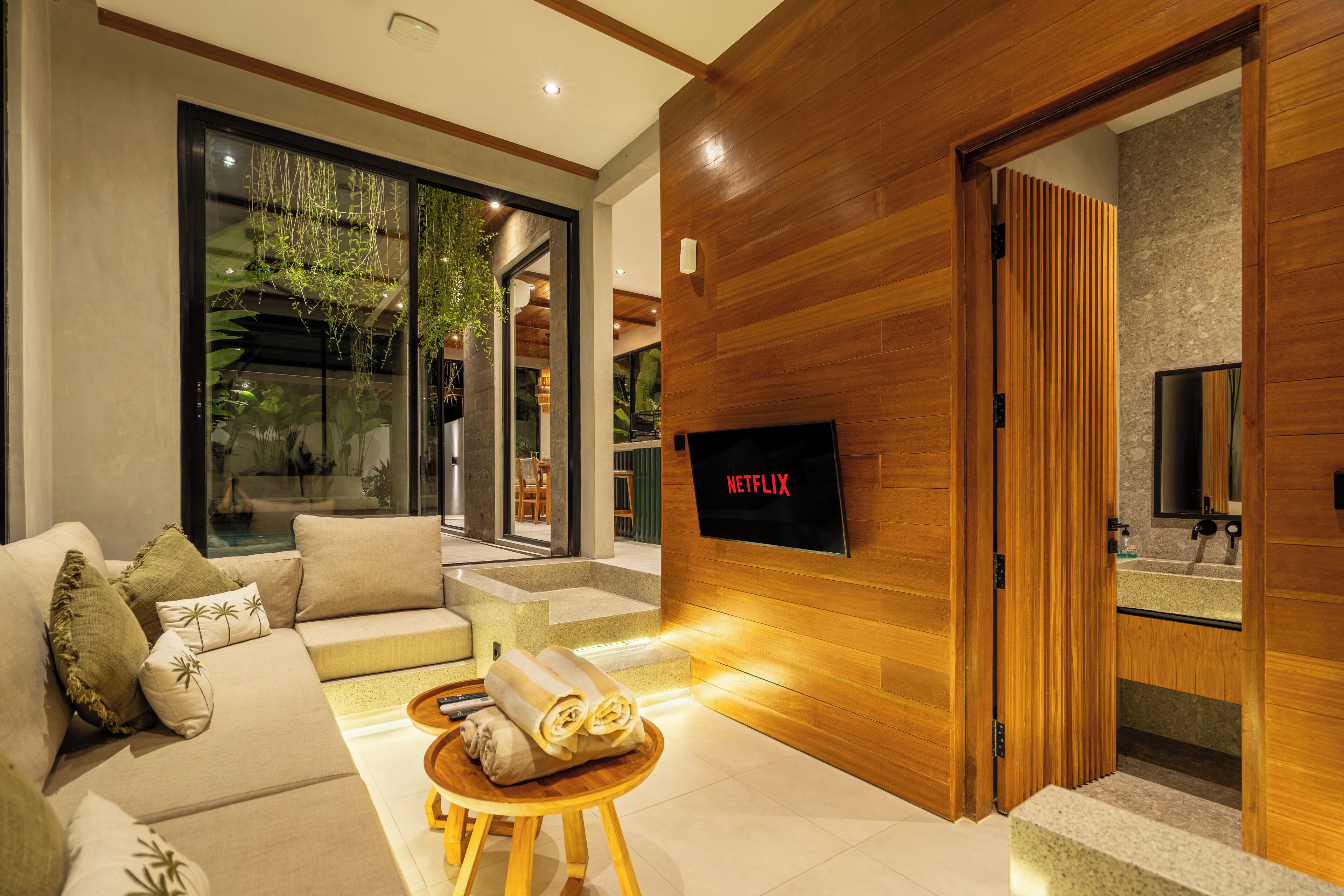 A modern living room with a sofa, a small wooden table with towels, a TV mounted on a wooden wall, and a glass door leading to a bathroom.