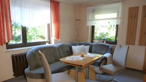 A room with a grey sofa, a wooden table, two chairs, a radiator, and windows with curtains.