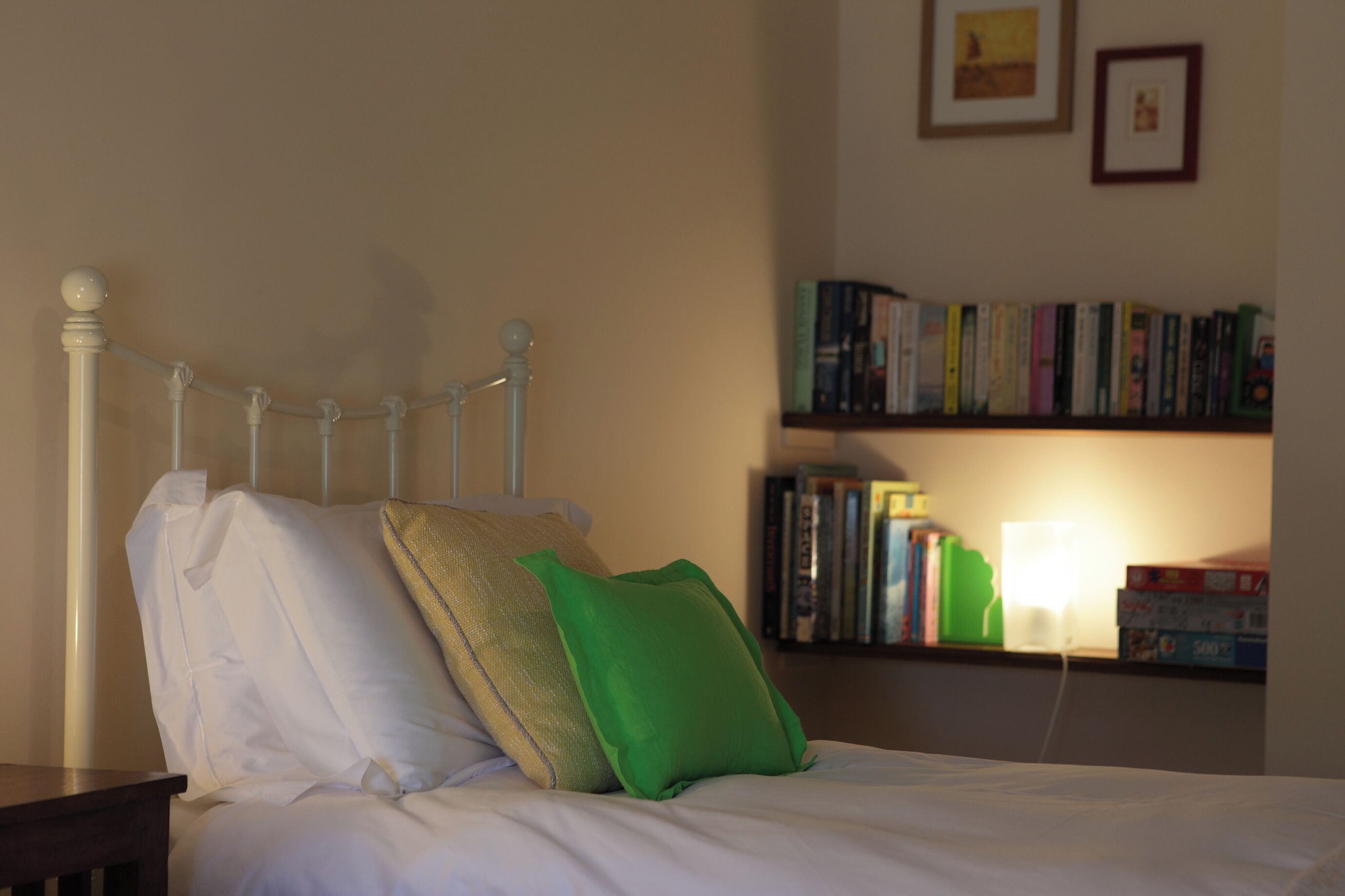 A bed with white and green pillows, a white metal headboard, and a bedside table. A bookshelf with books and a lamp is visible in the background.