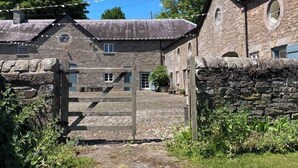 A stone building with a rustic entrance, a small garden, and a clear blue sky. - Glanhenwye Courtyard Cottages (Hereford)