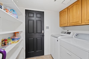 A laundry room with a black door, a washing machine, and shelves with laundry detergent. - Foothills Oasis Heated Pool&HotTub Stunning Views (Tucson)