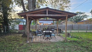 A covered patio with outdoor furniture, surrounded by a wooden fence and trees.