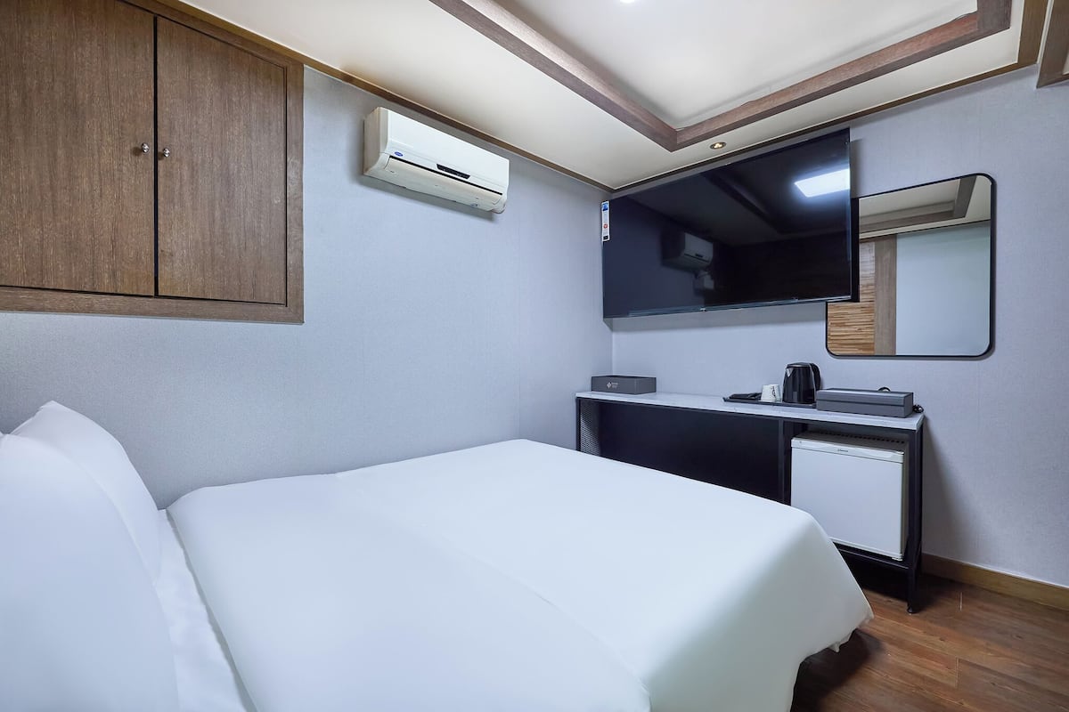 A hotel room with a bed, a desk, a mirror, an air conditioner, and a small refrigerator.