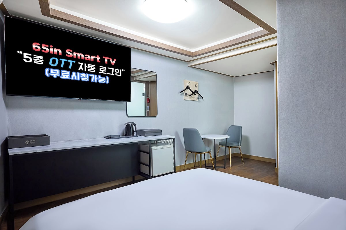 A modern hotel room with a large flat-screen TV, a desk with a kettle, a small fridge, and a bed.