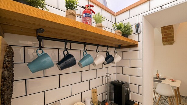 A kitchen with a wooden shelf holding plants and a fire extinguisher, a hanging pot rack with various pots, and a coffee machine.