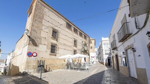 A narrow cobblestone street with a café setup on the left, featuring outdoor seating and umbrellas.