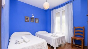 Une chambre avec deux lits, une chaise en bois, une fenêtre avec des rideaux et des tableaux accrochés au mur.