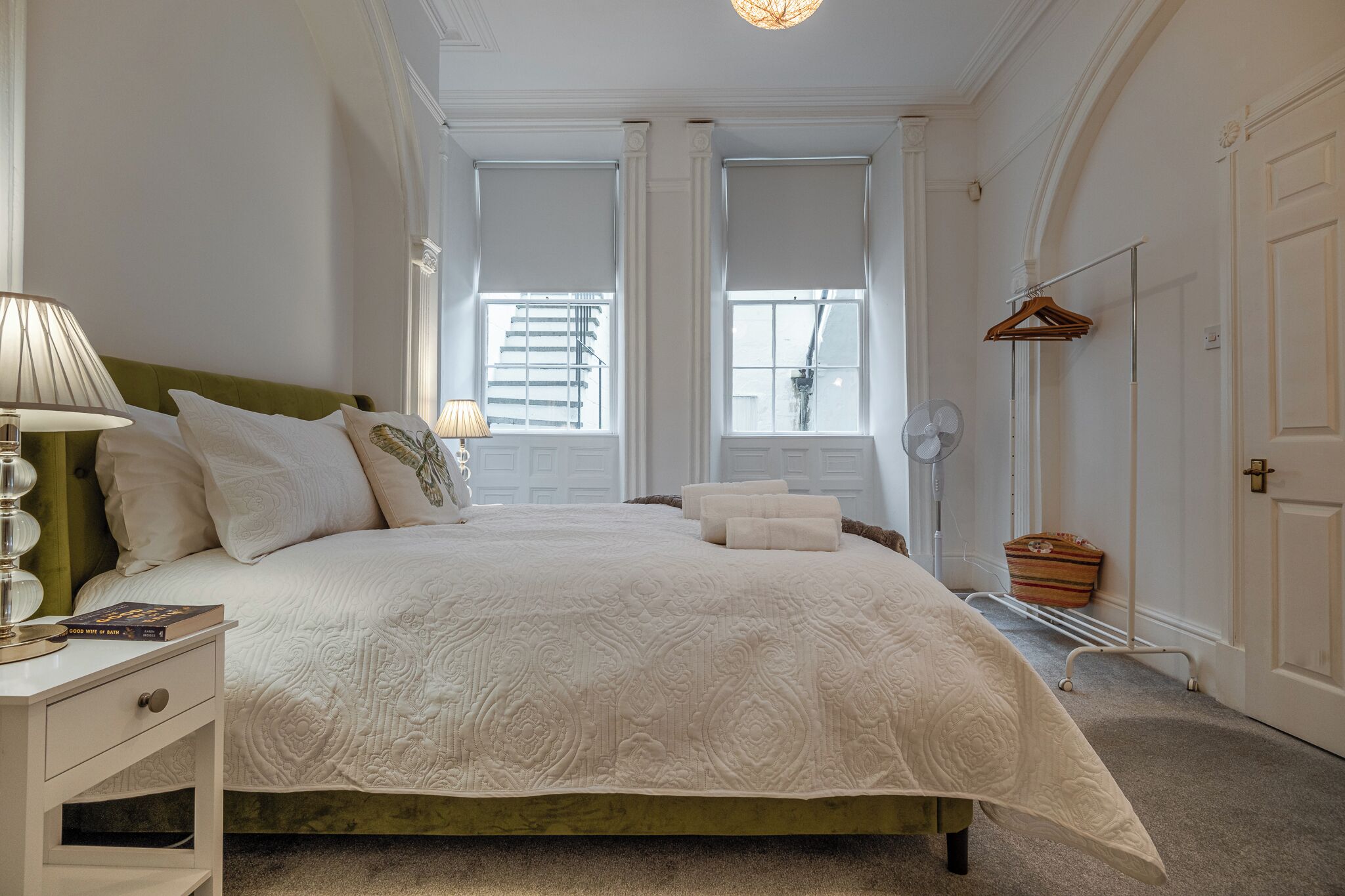 A neatly arranged bedroom with a large bed, bedside lamps, a bedside table, a hanging clothes rack, and a window with blinds.