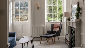 A cozy living room with a fireplace, a comfortable armchair, a small table, and a view of the outdoors through large windows. - Garden Flat Apartment in Grosvenor Place (Bath)