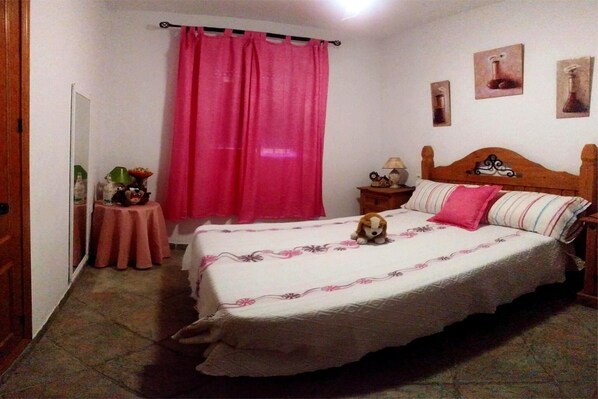 A bedroom with a bed, a nightstand, a mirror, a pink curtain, and a small table with a lamp.